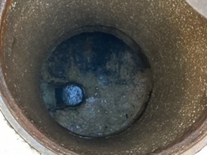 A dirty grease trap. The trap is a circular pit containing water and solidified fats, oils, and greases. The fats, oils, and greases are brown and floating on top of the water.