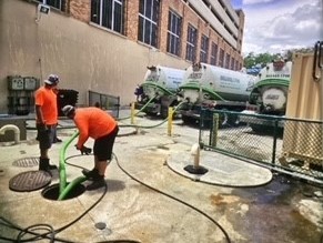 Two workers are pumping out a grease trap.  The grease trap is a hole in the sidewalk that 2 tubes are coming out of.  The 2 tubes are sucking the FOGs out of the trap and into 3 trucks parked behind the trap.