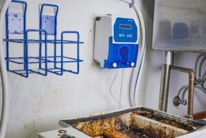 Blue chemical dosing pump mounted on wall next to dirty sink in commercial facility maintenance room