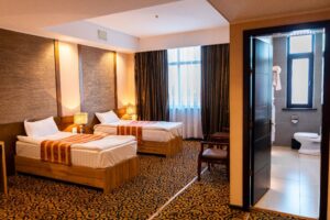 Modern hotel guest room with twin beds, dark curtains, patterned carpet, and attached glass bathroom