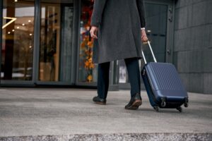 Business traveler pulling blue rolling suitcase on concrete sidewalk outside commercial building entrance